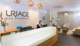 L'image présente un comptoir d'accueil de spa avec une personne derrière, dans un espace où prédominent le blanc, le bois clair et des touches de bleu.
