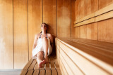 La photo montre l'intérieur d'un sauna en bois clair, avec une personne assise sur un banc.