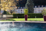 L'image présente un espace aquatique extérieur avec une piscine au premier plan. À l'arrière-plan, on distingue une haie de buissons touffus et, au-dessus, on aperçoit les bâtiments de l’avenue des thermes d’Uriage.