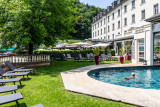 Une piscine extérieure-intérieure avec un homme se baignant. La piscine est entourée par un espace engazonné avec des transats gris. On aperçoit le grand hôtel et sa terrasse au fond.
