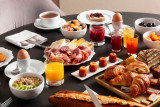 Sur une table sombre, un assortiment de mets sucrés et salés est disposé avec des viennoiseries, du pain, de la charcuterie, des œufs, des fruits frais, des boissons et des confitures.