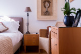 Cette chambre présente un lit double agrémenté de coussins et d'une couverture à motifs, une table de chevet en bois avec une lampe, et une commode assortie, le tout dans des tons clairs, avec une touche de verdure grâce à une orchidée.