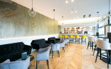Ce bar présente un espace en longueur avec un comptoir au fond et une série de tables avec des canapés le long du mur à gauche et des tables hautes à droite.