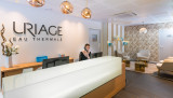 L'image présente un comptoir d'accueil de spa avec une personne derrière, dans un espace où prédominent le blanc, le bois clair et des touches de bleu.