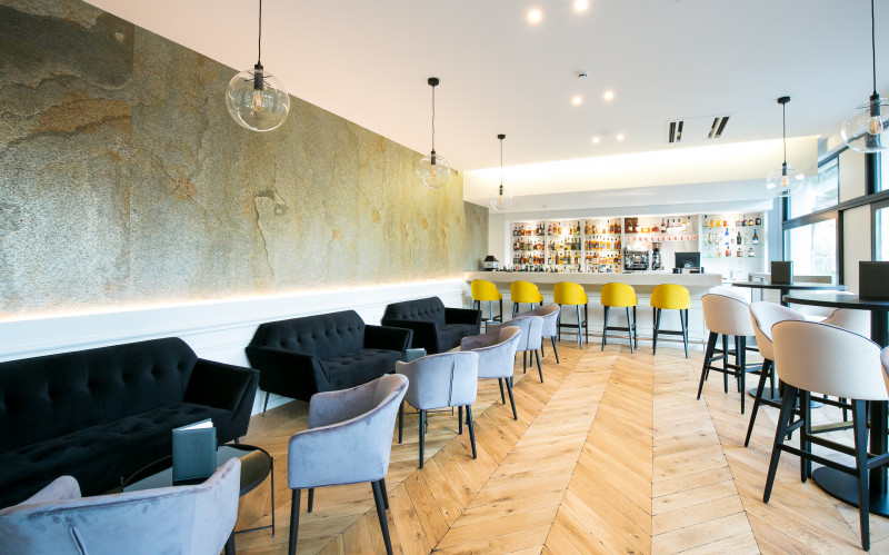 Ce bar présente un espace en longueur avec un comptoir au fond et une série de tables avec des canapés le long du mur à gauche et des tables hautes à droite.