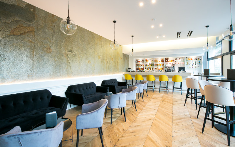 Ce bar présente un espace en longueur avec un comptoir au fond et une série de tables avec des canapés le long du mur à gauche et des tables hautes à droite.